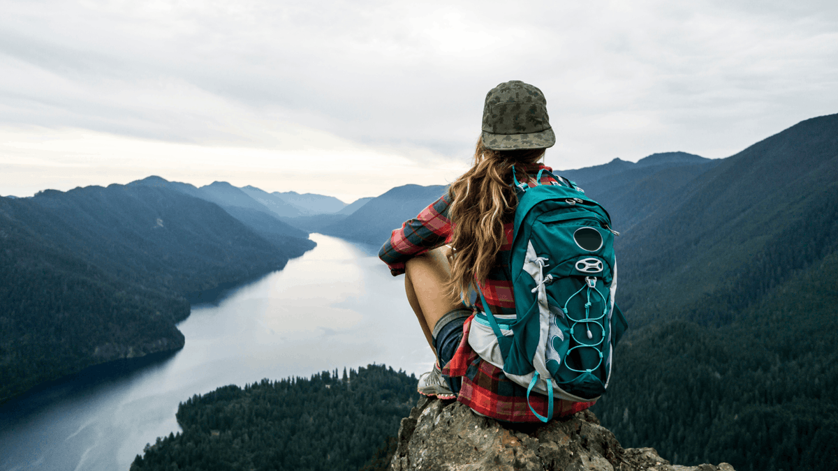 Person with a backpack, with long flowing brown hair wearing a camp hat backwards sitting on a mountain overlooking a lake and mountains.