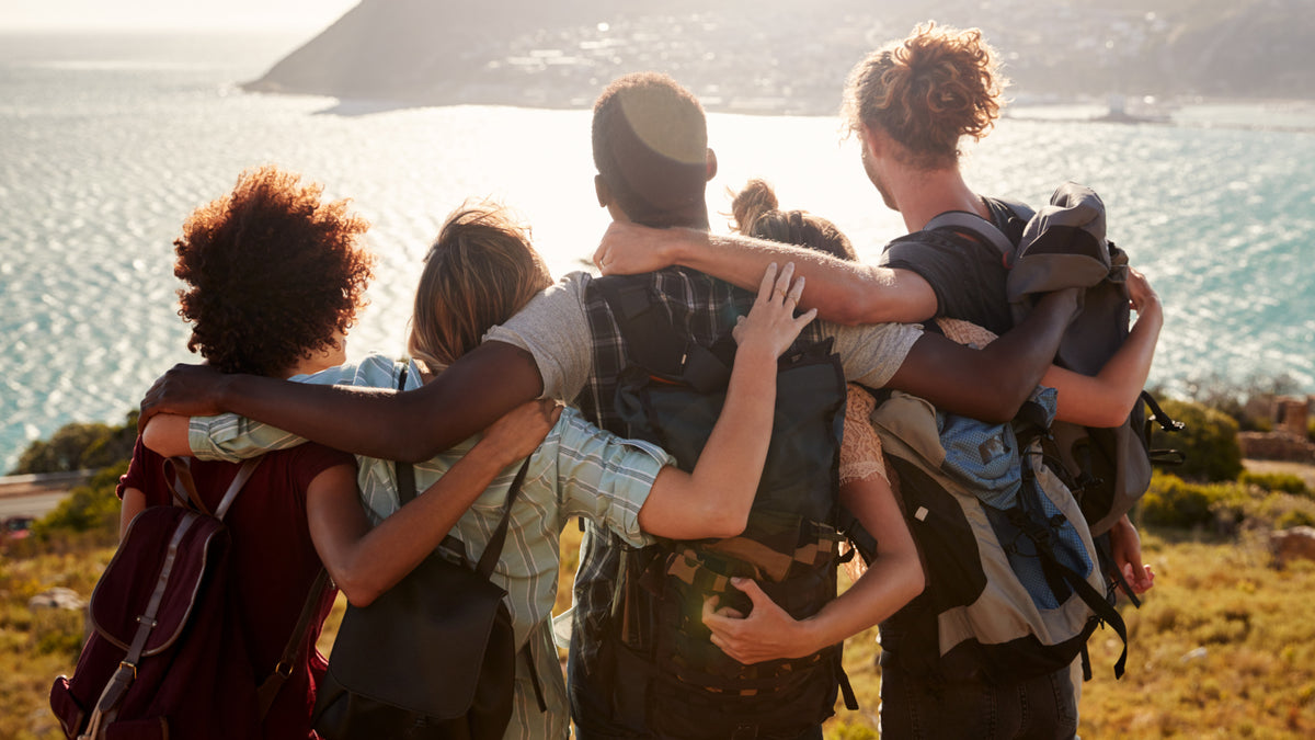 Five friends standing arm in arm on a hill overlooking the ocean and mountains all wearing backpacks 