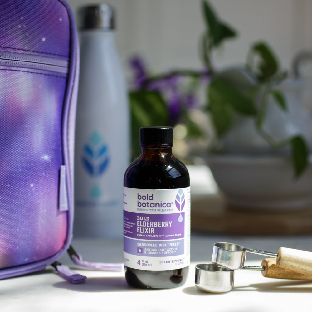 bottle of bold botanica bold elderberry elixir on a kitchen countertop sitting next to metal measuring spoons and a purple bag with a white metal water bottle sporting with blue bold botanica logo blurred in the background