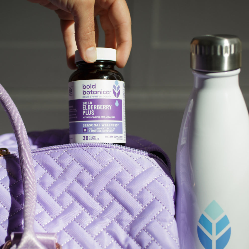 Bottle of bold botanica bold elderberry plus extract being put into a purple gym bag sitting next to a white metal water bottle sporting the blue bold botanica logo