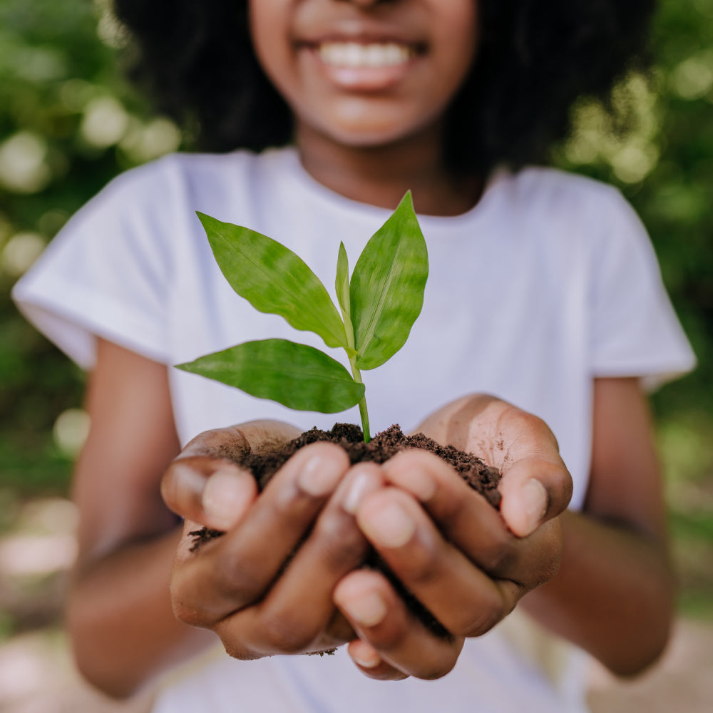 Young girl holding a tree sapling in her hands wearing a white t shirt and a blurry green tree background