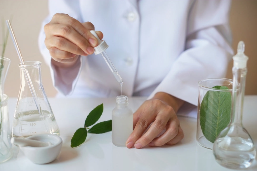 Lab photo with a dropper being raised, dispensing clear liquid into a bottle but a woman in a lab coat standing behind a white counter.  other glass beakers on the right and left of the photo with a leaf in one of the beaker on the right