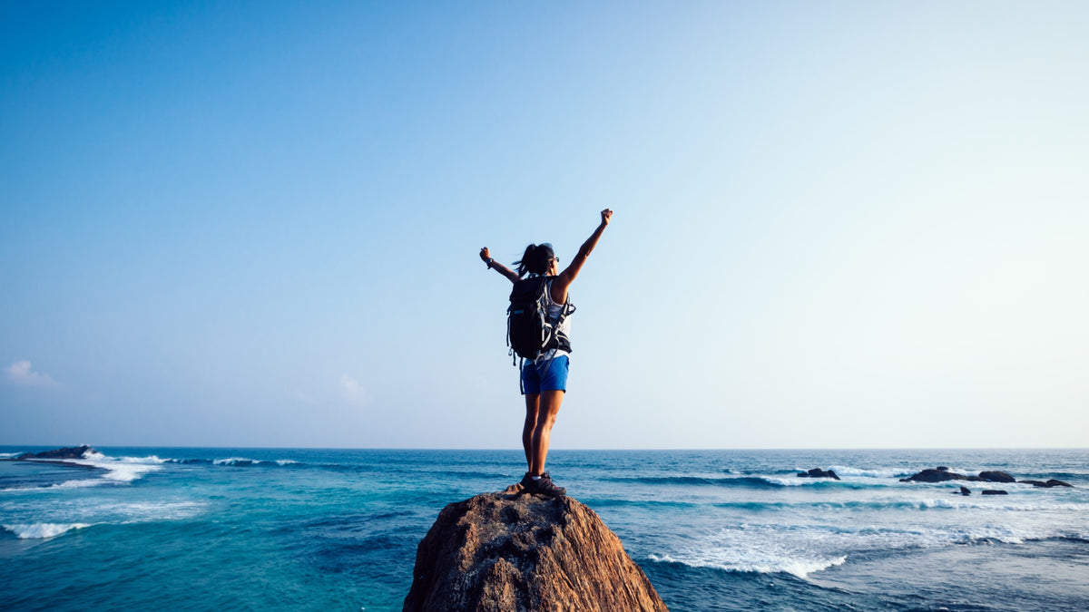 woman with arms raised wearing a backpack standing on rock in the ocean with a clear blue sky. 